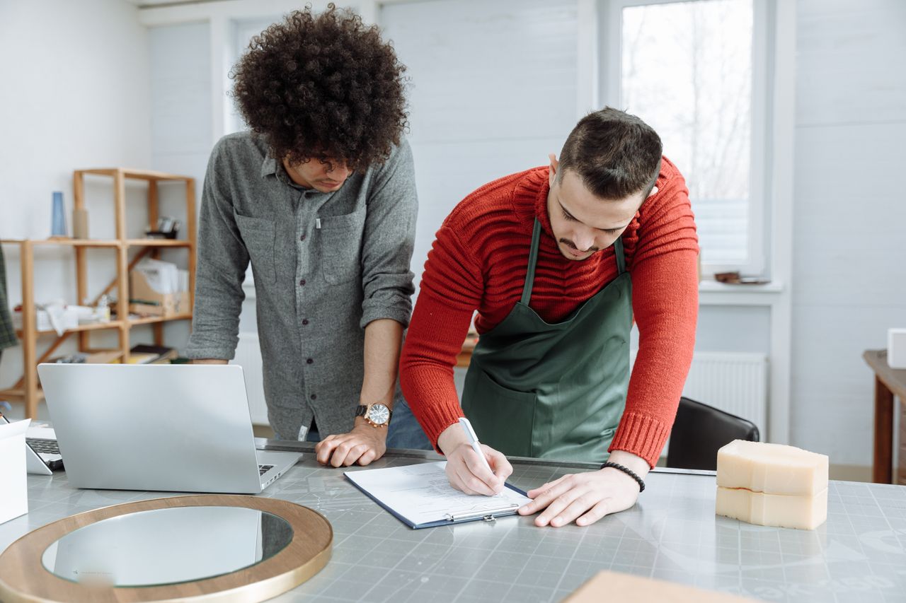 Two men working together at a desk, taking notes in a modern workshop environment.
